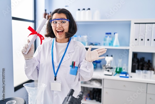 Young hispanic woman working at scientist laboratory holding degree celebrating achievement with happy smile and winner expression with raised hand