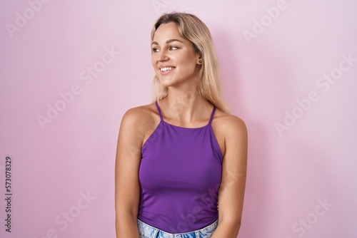 Young blonde woman standing over pink background looking away to side with smile on face, natural expression. laughing confident.