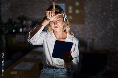 Young blonde woman working at the office at night making fun of people with fingers on forehead doing loser gesture mocking and insulting.