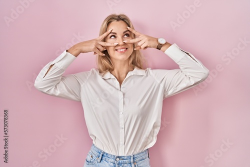 Young caucasian woman wearing casual white shirt over pink background doing peace symbol with fingers over face, smiling cheerful showing victory © Krakenimages.com