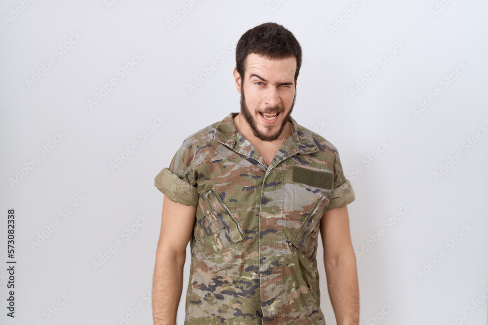 Young hispanic man wearing camouflage army uniform winking looking at the camera with sexy expression, cheerful and happy face.