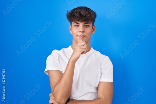Hispanic teenager standing over blue background with hand on chin thinking about question, pensive expression. smiling and thoughtful face. doubt concept.