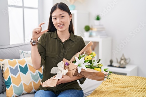 Chinese young woman holding bouquet of white flowers smiling and confident gesturing with hand doing small size sign with fingers looking and the camera. measure concept.