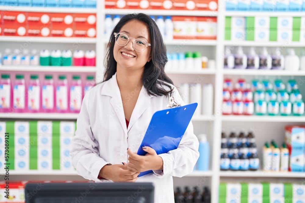 Asian young woman working at pharmacy drugstore holding clipboard looking positive and happy standing and smiling with a confident smile showing teeth