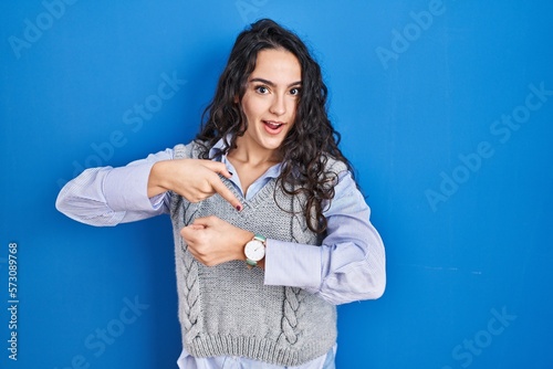 Young brunette woman standing over blue background in hurry pointing to watch time, impatience, upset and angry for deadline delay