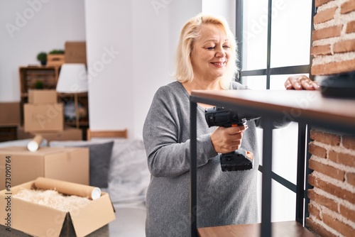 Middle age blonde woman smiling confident repairing shelving at new home