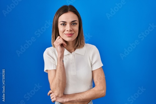 Hispanic woman standing over blue background looking confident at the camera smiling with crossed arms and hand raised on chin. thinking positive.