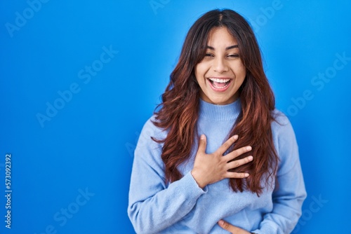 Hispanic young woman standing over blue background smiling and laughing hard out loud because funny crazy joke with hands on body.