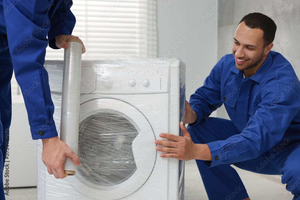 Obraz premium Male movers with stretch film wrapping washing machine in bathroom. New house