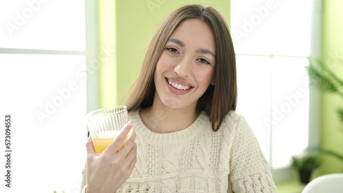 Young beautiful hispanic woman holding glass of orange juice sitting on table at home