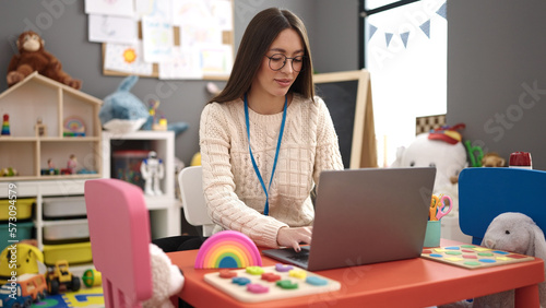 Young beautiful hispanic woman preschool teacher using laptop sitting on table at kindergarten