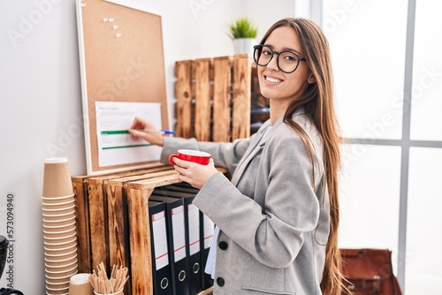 Young beautiful hispanic woman business worker drinking coffee writing on cork board at office