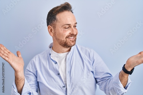 Middle age caucasian man standing over blue background smiling showing both hands open palms, presenting and advertising comparison and balance © Krakenimages.com