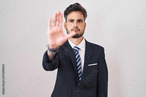 Young hispanic man with tattoos wearing business suit and tie doing stop sing with palm of the hand. warning expression with negative and serious gesture on the face. © Krakenimages.com