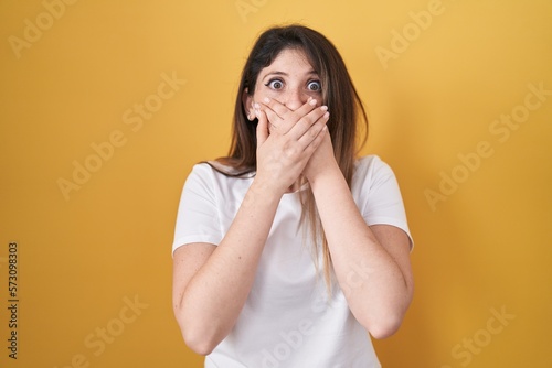 Young brunette woman standing over yellow background shocked covering mouth with hands for mistake. secret concept.