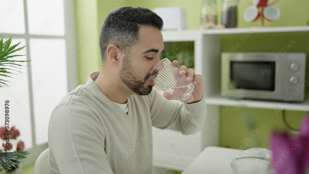 Young hispanic man drinking water sitting on table at home