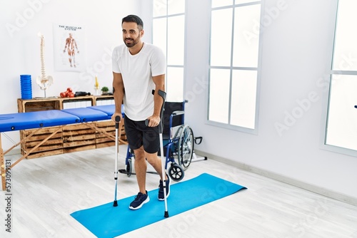 Young hispanic man patient having rehab session walking using crutches at clinic photo