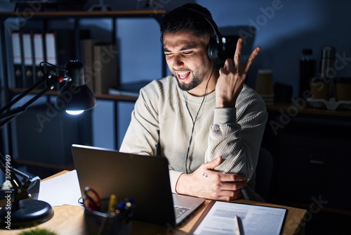 Young handsome man working using computer laptop at night smiling with happy face winking at the camera doing victory sign. number two.