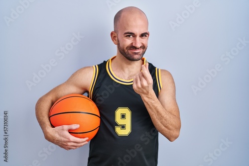 Young bald man with beard wearing basketball uniform holding ball doing money gesture with hands, asking for salary payment, millionaire business