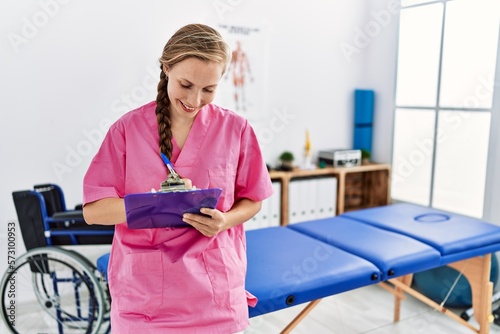 Young caucasian woman wearing physiotherapist uniform writing on checklist standing at physiotherapy clinic