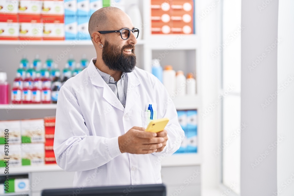 Young bald man pharmacist using smartphone working at pharmacy