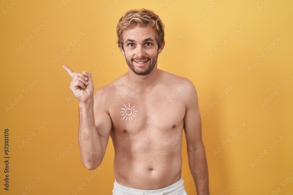 Caucasian man standing shirtless wearing sun screen with a big smile on face, pointing with hand finger to the side looking at the camera.