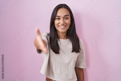 Young hispanic woman standing over pink background smiling friendly offering handshake as greeting and welcoming. successful business.
