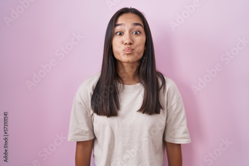 Young hispanic woman standing over pink background puffing cheeks with funny face. mouth inflated with air, crazy expression.