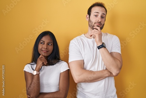 Interracial couple standing over yellow background with hand on chin thinking about question, pensive expression. smiling and thoughtful face. doubt concept.