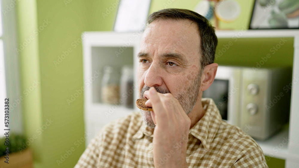 Middle age man eating cookie sitting on table at home