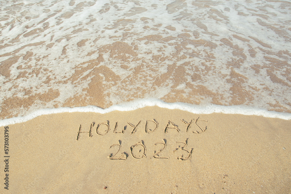 Fototapeta premium Holidays 2023 lettering on the beach with wave and clear blue sea.