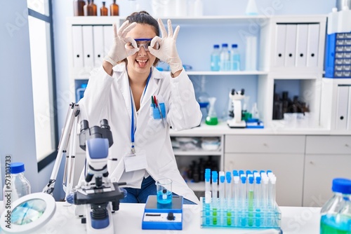 Young hispanic woman working at scientist laboratory doing ok gesture like binoculars sticking tongue out, eyes looking through fingers. crazy expression.