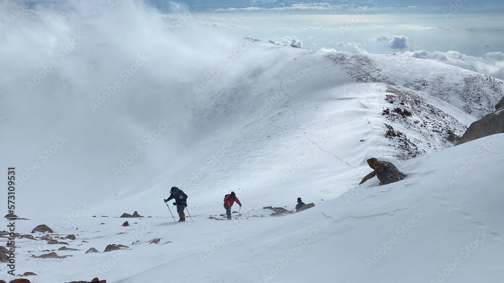 Climbers with small backpacks and trekking poles are walking on a snowy slope. Walk above the clouds. A group of tourists climbs uphill in a heavy snowstorm. Climbers with small backpacks and trekking poles are walking on a snowy slope. Walk above the clouds. A group of tourists climbs uphill in a heavy snowstorm.