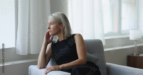 Middle age thoughtful, pensive woman sits on armchair staring out of window, ponders, thinking, missing or waiting, looks frustrated, recall life and recollects memories, resting alone in living room photo