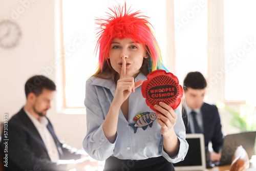 Young woman with funny wig and whoopee cushion showing silence gesture in office. April Fools' Day celebration photo