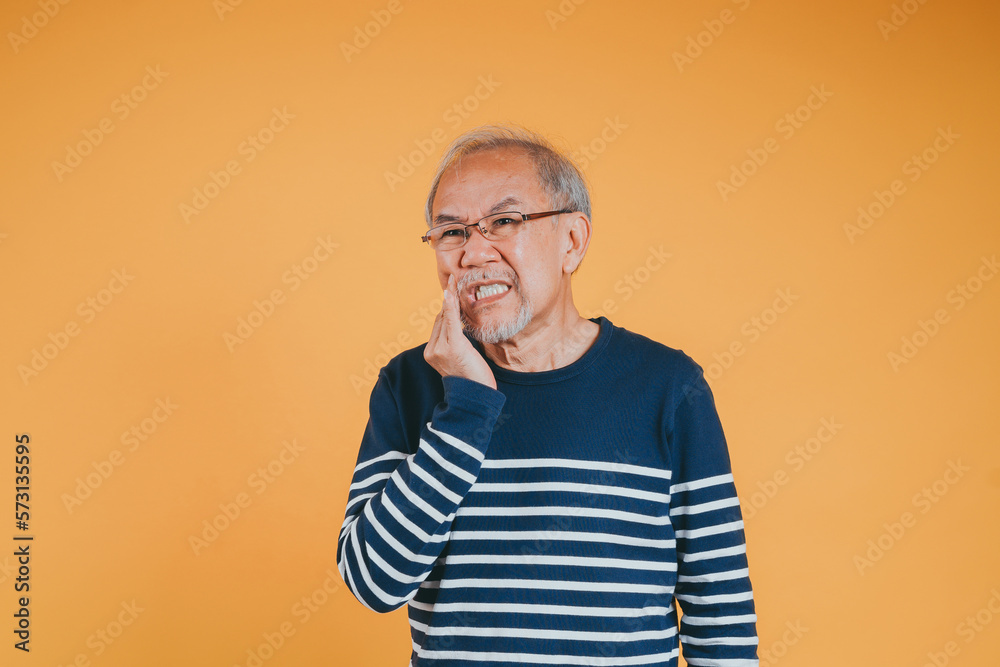 Fototapeta premium Dental pain. Portrait senior old man sad hand touching cheek suffering from toothache studio shot isolated on yellow background, Asian unhappy elder man problems with teeth pain, dental healthcare