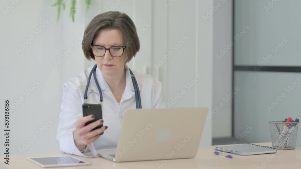 Old Female Doctor using Smartphone while using Laptop Old Female Doctor using Smartphone while using Laptop