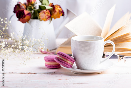 French macaroon dessert. Bright pink macaroons and a white cup of coffee on a white wooden table against the backdrop of an open book and a winter bouquet of dried flowers