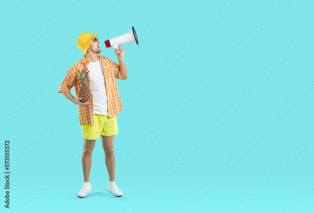 Young man wearing summer shirt, shorts, sun hat and sunglasses standing on blank turquoise copyspace studio background, holding pineapple and megaphone and making announcement about great holiday sale Young man wearing summer shirt, shorts, sun hat and sunglasses standing on blank turquoise copyspace studio background, holding pineapple and megaphone and making announcement about great holiday sale