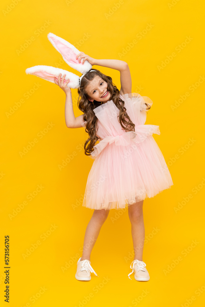 Easter children's holiday. A teenage girl in a lush dress rejoices in spring. The child is wearing long rabbit ears. A beautiful little girl with long hair, full-length on a yellow isolated background Easter children's holiday. A teenage girl in a lush dress rejoices in spring. The child is wearing long rabbit ears. A beautiful little girl with long hair, full-length on a yellow isolated background