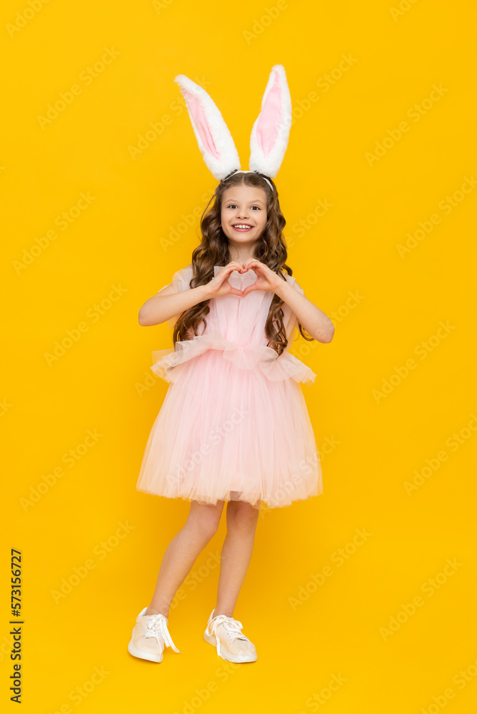 Festive Easter. A teenage girl in a fluffy dress and with rabbit ears smiles broadly and shows her heart. A beautiful little girl in a rabbit costume on a yellow isolated background, full-length. Festive Easter. A teenage girl in a fluffy dress and with rabbit ears smiles broadly and shows her heart. A beautiful little girl in a rabbit costume on a yellow isolated background, full-length.