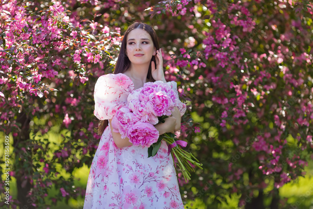 A pretty brunette in a dress, with a large bouquet of peonies, stands near pink blooming apple trees A pretty brunette in a dress, with a large bouquet of peonies, stands near pink blooming apple trees
