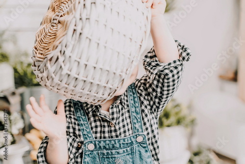 Little girl 1 years old with a basket on her head. Funny child plays puts a basket on her head like a hat and laughs, she is dressed in a plaid shirt and denim overalls. Funny moments with kids. © svetograph