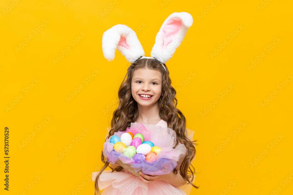 Festive Easter for children. A beautiful teenage girl with curly hair holds a basket of colored colored eggs for the holiday. A child in a rabbit costume with long ears on a yellow isolated background