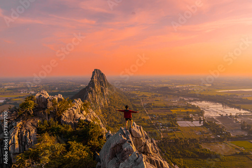 Picture from the back of a man standing on the peak of high mountain cliff. There are a lot of rock with white color. The sun is setting on the mountain and there is a beautiful warm orange light. © Pang wrp