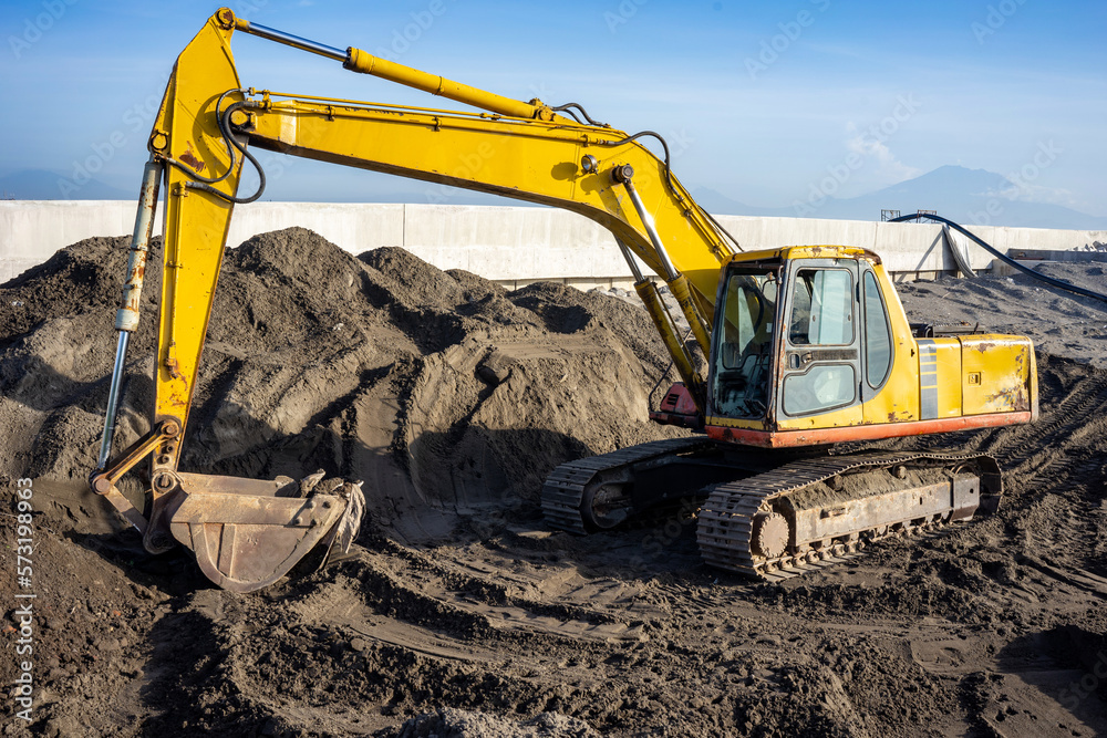 A yellow big excavator stands on a construction site with dark soil on a sunny day with a blue sky. A yellow big excavator stands on a construction site with dark soil on a sunny day with a blue sky.