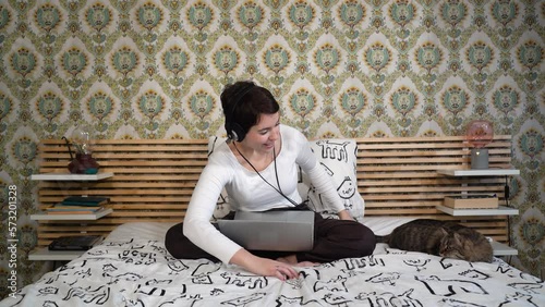 The beautiful woman is relaxing on the bed with her cat. The woman is lying down and she plays with the gray cat while she listens to music.
