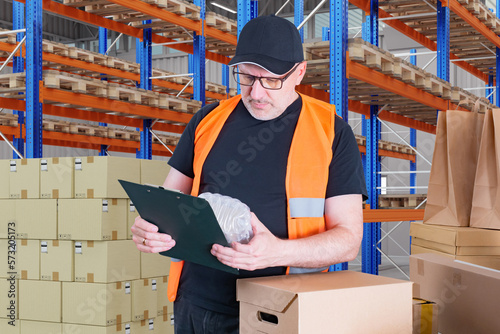 Man with clipboard in warehouse. Supervisor controls acceptance of goods. Guy in orange vest works in warehouse. Man stands among boxes and racks. Supervisor conducts warehouse audit. photo