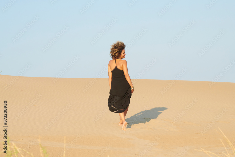 Anonymous woman in dress walking on beach. Back view of alluring ethnic woman in black dress walking on sandy coast in bright sunlight