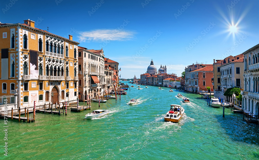 Grand canal in Venice with old houses, boats, the church Santa Maria della Salute in the background and sunburst in the blue sunny sky.